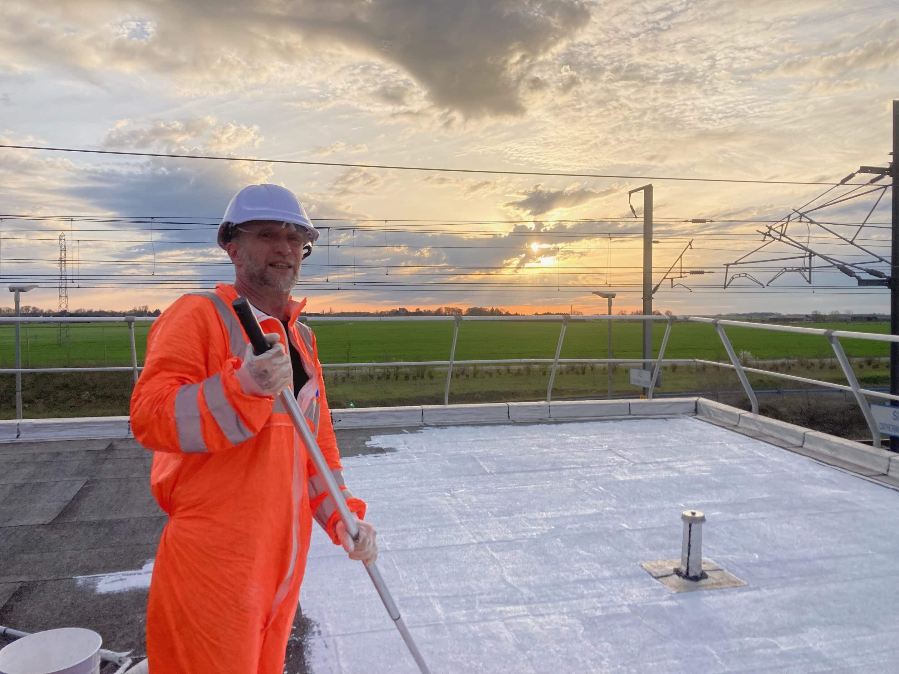 Un homme en combinaison orange et casque peignant un toit avec un rouleau sous un ciel nuageux.